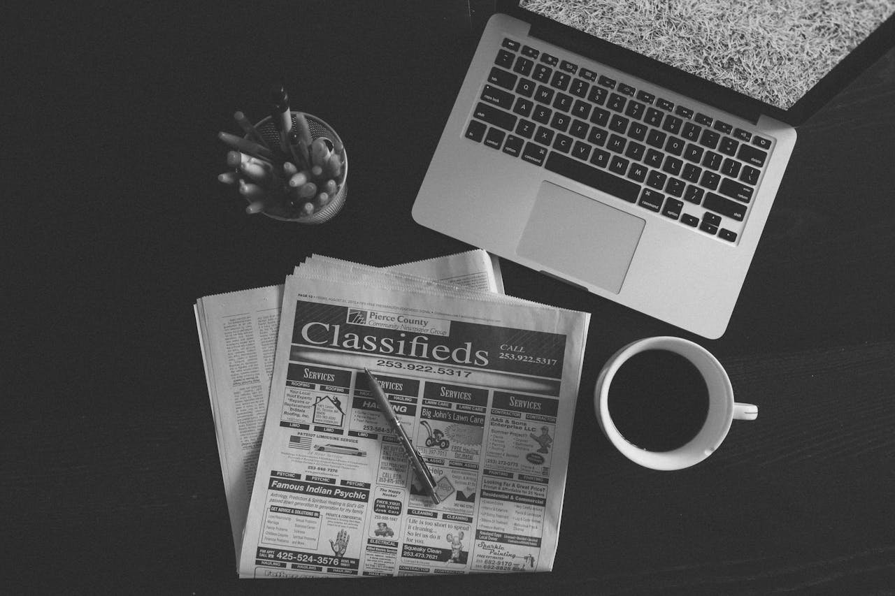 our-story A black and white image of a laptop, newspaper, and coffee cup on a desk.